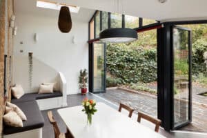 A modern dining area with a white table, chairs, and a vase of flowers. Large glass doors open to a patio with greenery and a stunning glazed gable end. Features include a skylight and bench seating with cushions, blending elegance with comfort seamlessly.
