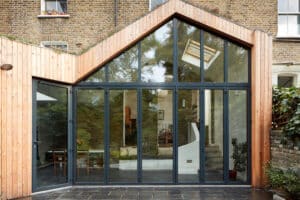 A modern house extension with a striking glazed gable end, featuring large glass doors and elegant wooden cladding, seen from the outside.