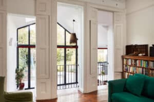 A modern living room features large windows and a glazed gable end, accentuating the white walls and green sofa. A pendant light elegantly hangs from the ceiling, while a bookshelf adds charm. The garden view through the expansive glass completes this harmonious space.