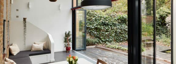 Modern interior with large glass doors and a stunning glazed gable end opening to a garden. Inside, there's a seating area with cushions and a white table, surrounded by ivy-covered walls and stone flooring outside.