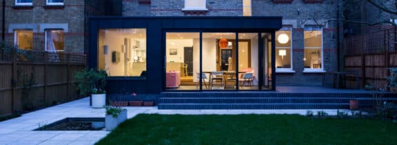 Modern house extension with large picture windows, showcasing a well-lit interior. The garden features a lawn and tiled patio area with potted plants.