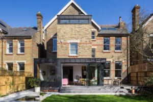 A modern two-story brick house with large picture windows, a glass-walled extension, and a well-maintained backyard under a clear blue sky.