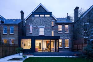 A two-story brick house with large picture windows, a modern glass extension, and a landscaped backyard beautifully illuminated at dusk.