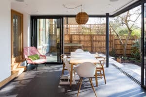 Modern dining area featuring a wooden table, a mix of chairs, and a pink lounge chair. A large picture window and glass doors open up to a fenced garden, allowing natural light to flood the space. A wooden pendant light adds warmth and elegance.