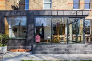 Modern house with large glass sliding doors and a stepped entrance. A picture window inside frames the view, enhancing the dining area with chairs and a pink armchair. Reflections of trees dance across the glass, creating an inviting and harmonious space.