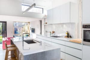 Modern kitchen with a marble island, white cabinets, and stainless steel appliances. Large picture windows provide natural light, offering a stunning view of the garden visible through open glass doors.