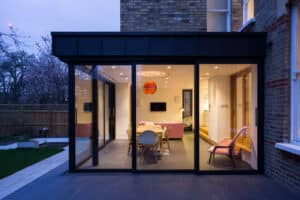 A modern glass-walled extension of a house features a dining area, pink sofas, and a lit orange pendant light, all framed beautifully by a large picture window. The scene is bathed in the serene glow of dusk, offering an inviting glimpse from outside.