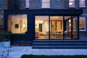 Glass-walled modern extension with dining area and a striking picture window. The illuminated interior reveals a visible kitchen, while the exterior showcases brickwork and potted plants, opening onto a landscaped patio with grass.