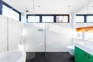 Modern bathroom with white subway tiles, a glass shower partition, black floor tiles, a green cabinet, and a round bathtub. Ambient light streams through high picture windows.