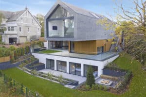Modern multi-level house with a sloped roof, large glass windows, and a spacious terrace surrounded by greenery.