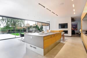 Modern kitchen with a large island, stools, and pendant lights. Floor-to-ceiling windows and bifold sliding doors offer a garden view. Minimalist design with neutral colors.