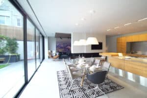 Modern open-plan kitchen and dining area with large glass doors, white walls, wooden cabinets, and a black and white patterned rug. Cozy seating area visible in the background.