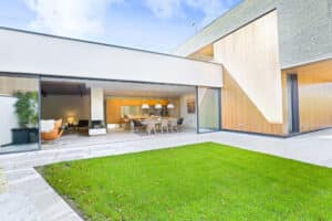 Modern house with large sliding glass doors opening to a patio and garden. Inside, there is a dining table, chairs, and a potted plant. Walls are wood-paneled and brick.