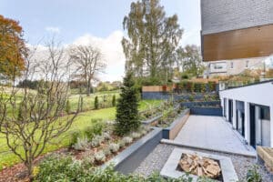 Modern terrace with neatly arranged garden beds and a fire pit, overlooking a green landscape with trees under a partly cloudy sky.