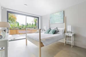 Modern bedroom with a large bed, two side tables, and a lamp. A large window offers a view of a garden. Walls are painted white and there's art above the bed.