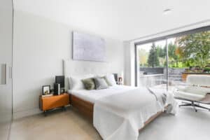 Modern bedroom with a large bed, wooden nightstands, cushioned chair, and a large window overlooking a garden. Walls are white, with minimalist decor and a light-colored abstract painting.