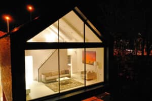 A modern house with large glass windows is illuminated at night. Inside, a sofa, rug, and a colorful painting are visible in the living room.