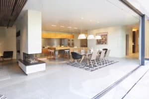Modern open-plan kitchen and dining area with wood accents, a fireplace, and large glass windows, featuring a long dining table surrounded by chairs and overhead pendant lights.