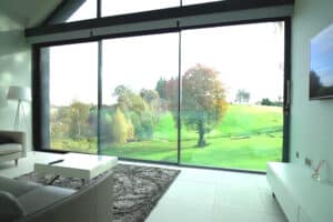 A modern living room with a large window overlooking a green golf course and trees. A sofa, coffee table, and TV are in view.