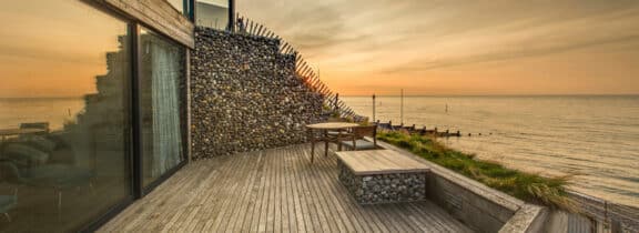 A wooden deck with an elegant stone wall overlooks the ocean at sunset. A small table and bench are placed on the deck, surrounded by sleek aluminium sliding doors on the left side.