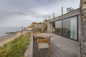 Beachfront patio with wooden furniture, overlooking the sea. A house with large glass doors and sleek aluminium sliding doors is on the right, and grass grows along the edge of the deck. Houses and a cloudy sky are in the background.