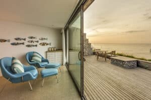 Modern living room with blue chairs and fish wall decor, featuring aluminium sliding doors that open to a wooden deck overlooking the ocean at sunset.