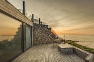 Modern coastal house with a glass facade, aluminium sliding doors, and stone accents, overlooking a wooden deck with a table and benches, facing a calm sea at sunset.
