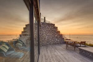 A seaside patio with wooden decking, lounge chairs, a stone wall, and a glass house exterior featuring sleek aluminium sliding doors. The scene overlooks a calm ocean at sunset.
