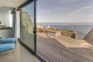 A room with sleek aluminium sliding doors opens to a wooden deck complete with chairs and a table, offering an unobstructed view of the ocean.