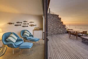 Modern living room with blue chairs and pillows, fish wall décor, and sleek aluminium sliding doors leading to an adjacent wooden deck overlooking the ocean at sunset.