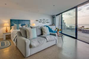 Modern bedroom with a double bed, sofa, and a breathtaking sea view through large aluminium sliding doors. Room decor includes fish-themed wall art and a small side table adorned with vibrant sunflowers, creating a serene coastal ambiance.