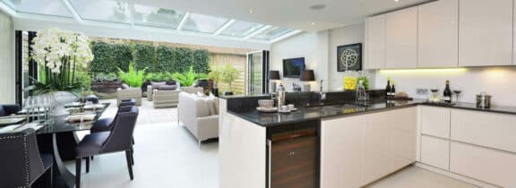 Modern kitchen and dining area with white cabinets, black countertops, and a glass ceiling. A sleek bifold door track seamlessly connects the indoor space to an outdoor patio with seating and lush greenery.