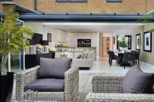 Spacious open-plan patio area with wicker chairs, seamlessly connecting to the modern kitchen and dining room through a bifold door track. Neutral tones and natural lighting create an inviting ambiance.