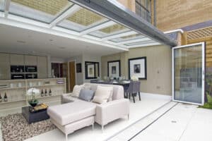 Modern open-concept living area with a kitchen, dining table, and a beige sectional sofa. Glass doors on a sleek bifold door track open to the patio, and the space features minimalist decor with framed wall art.
