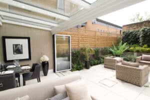A modern indoor-outdoor space featuring stylish wicker furniture and a glass ceiling, seamlessly connected by a bifold door track. The dining area opens to a patio bordered by a wooden fence and lush plants.