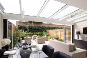 A modern indoor-outdoor space with a glass ceiling features a sleek bifold door track. In the foreground, a dining table sets the scene, while plush sofas invite relaxation. Beyond, a patio with chairs and lush greenery creates a seamless blend of nature and comfort.