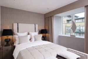 A modern bedroom with a beige color scheme, featuring a bed adorned with multiple pillows, two black lamps on either side, and a window framed by bifold door tracks offering a glimpse of neighboring buildings.