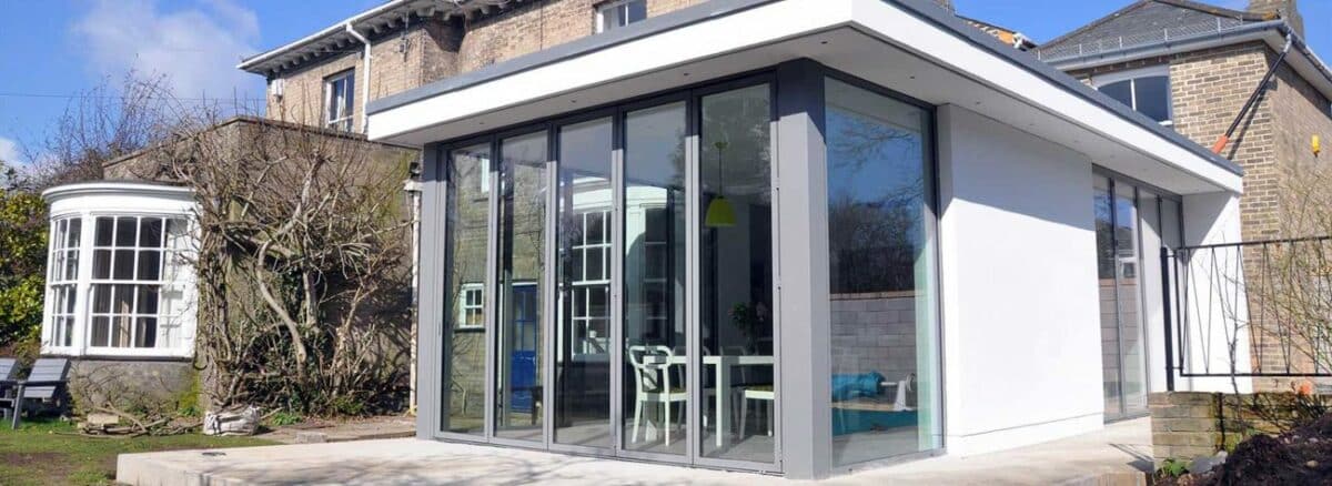 Modern glass extension with a flat roof and overhang seamlessly complements the older brick house, featuring sliding doors that open to a stunning garden view.