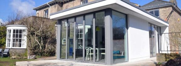 Modern glass extension with a flat roof and overhang seamlessly complements the older brick house, featuring sliding doors that open to a stunning garden view.