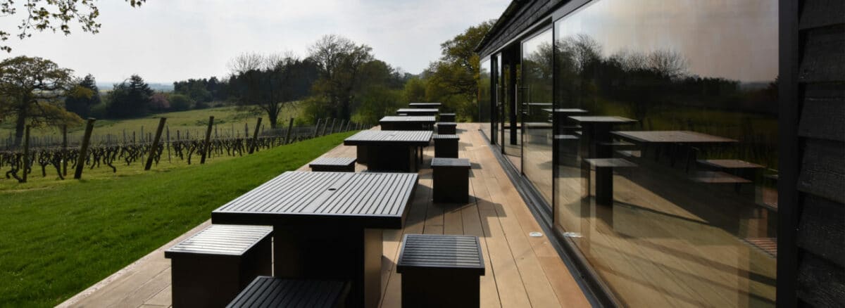 Outdoor terrace with black tables and benches overlooking a vineyard, adjacent to a glass-walled building featuring sleek aluminium sliding doors.