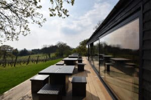Outdoor seating area with wooden tables and benches next to a building featuring large windows and sleek aluminium sliding doors, overlooking a grassy field and vineyard.