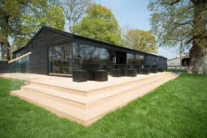 A modern black building with large glass windows and sleek aluminium sliding doors is surrounded by trees. It features a stepped wooden deck with multiple black tables and chairs on a grassy lawn, creating a seamless blend of nature and contemporary design.