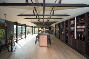 Modern wine tasting room with floor-to-ceiling windows and sleek aluminium sliding doors, wooden floors, a central bar with stools, and wall-mounted shelves filled with wine bottles.