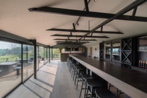 A modern bar with a long black counter and high stools, featuring sleek aluminum sliding doors. Large windows on the left provide natural light and a view of the outdoors.