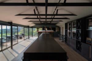 The long room, adorned with wooden beams and dark wood furniture, features large windows complemented by sleek aluminium sliding doors. Wine racks line the walls, offering a perfect view of the lush greenery outside.