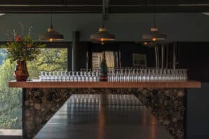 A bar counter with a row of empty wine glasses, a solitary bottle, and a floral arrangement is warmly illuminated by hanging lights. Behind it, aluminium sliding doors add a modern touch. The phrase "Great minds drink alike" is etched on the counter.
