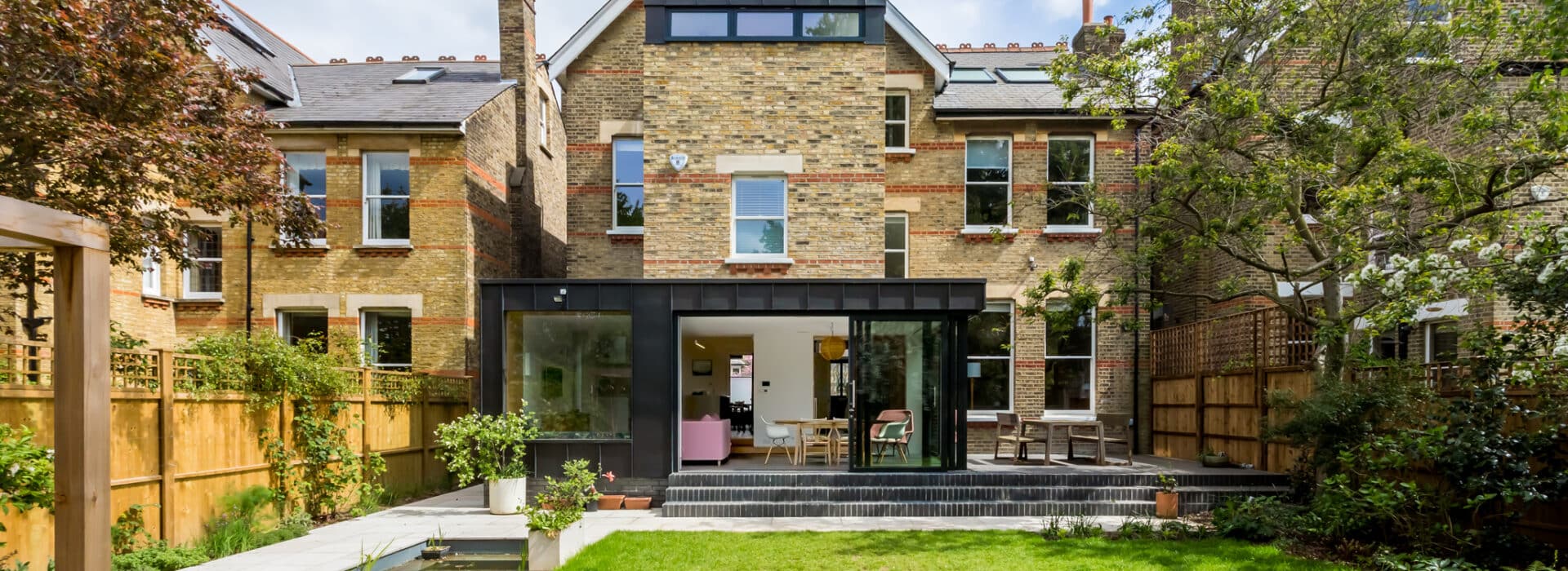 Modern extension on a traditional brick house, featuring a large picture window and glass doors opening to a patio and lawn, with outdoor seating and garden landscaping.