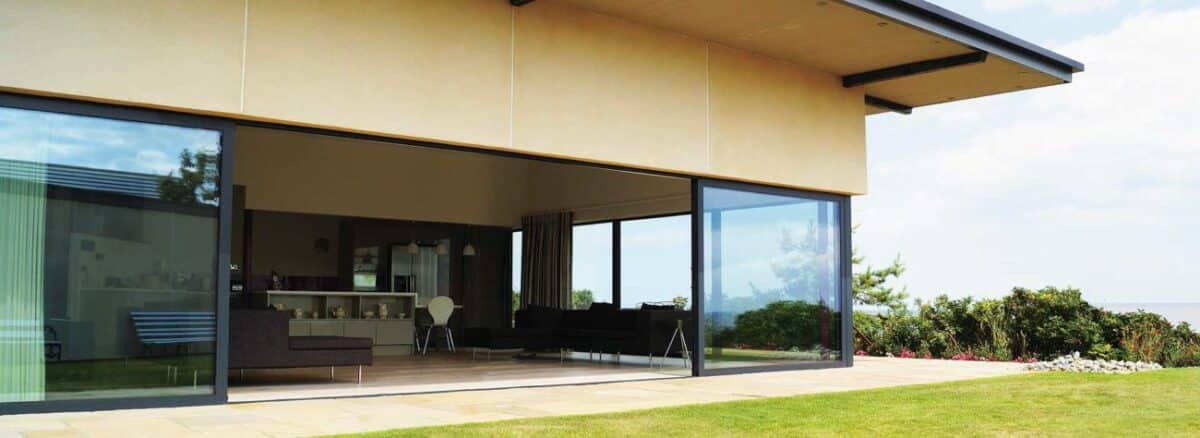 Modern house with large sliding glass doors and picture windows, overlooking a well-maintained lawn and garden, offering a glimpse of the interior living space.