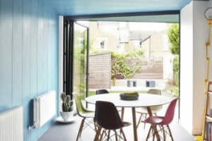 Modern dining room with a round table, colorful chairs, and a light wood shelf. A large open window with bifold doors reveals a small patio with a wooden fence and greenery. Blue walls and ceiling add to the serene charm.