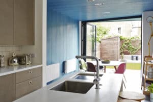 Modern kitchen with a white countertop, sink, and appliances. Blue-paneled open wall with bifold doors reveals a patio with table, chairs, and garden. Bright, natural light fills the space.